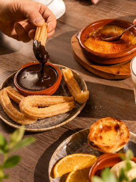 Churros mit Zimt-Zucker, die in dicke Schokoladensauce getaucht werden, daneben Crème Catalana und Kaffee.