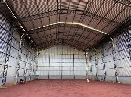 Vista interna de quadra tênis coberta com estrutura metálica, fechamento em telhas e piso de saibro.