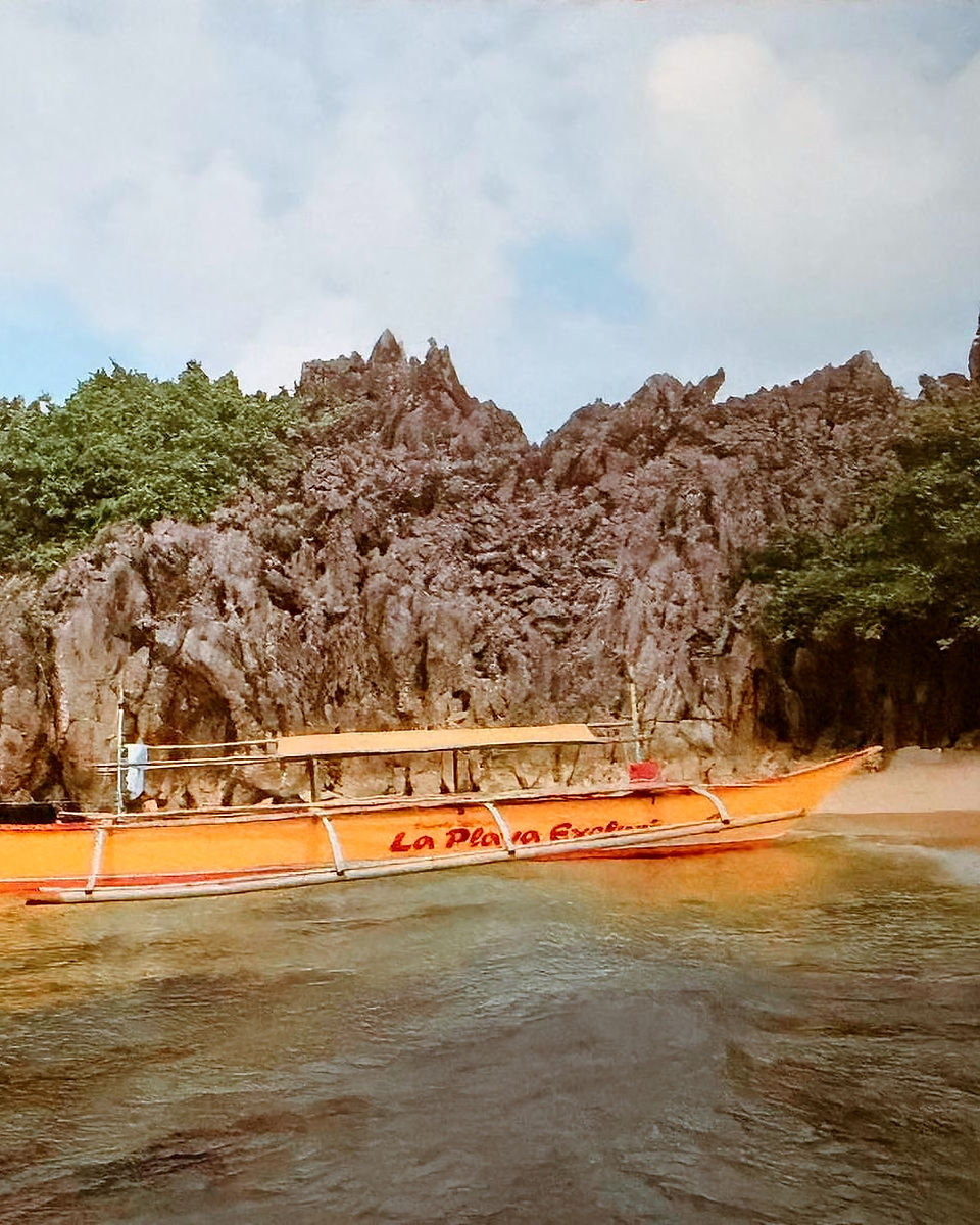 Photographie de 2010 montrant une banca traditionnelle philippine nommée La Playa accostant sur la plage de sable blanc de Lahus Island, encadrée par de hautes falaises de calcaire karstique dans l'archipel de Caramoan
