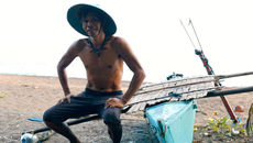 Un pêcheur local, arborant son fusil de pêche, se tient fièrement à côté de sa banca traditionnelle (pirogue à balancier) sur une plage de sable noir de Camiguin, Philippines