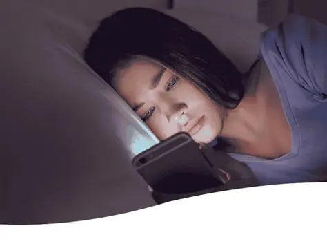 Kind liegt im Bett in einer dunklen Umgebung und schaut auf ihr Handy welches hell in das Gesicht scheint