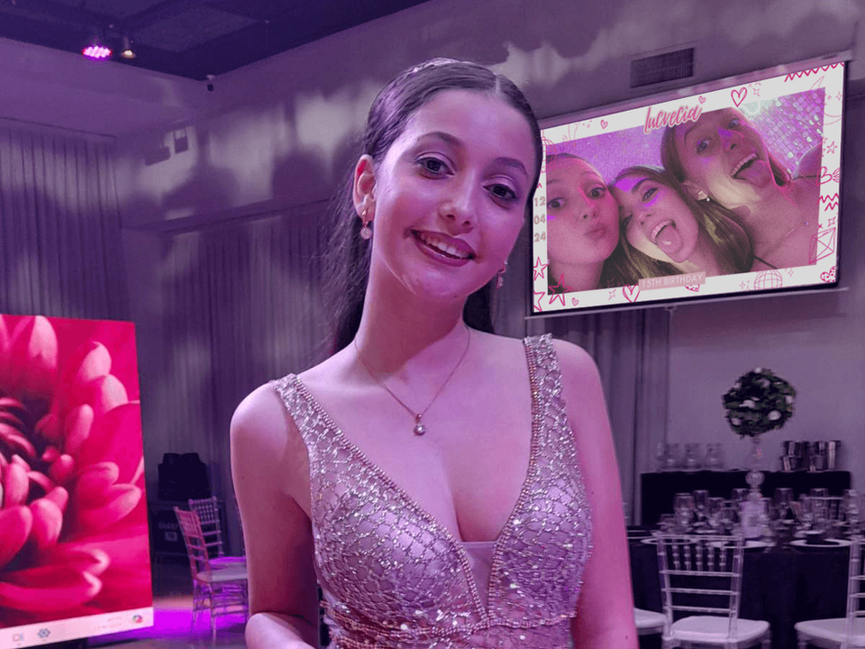 Hermosa quinceañera posando para una foto durante su fiesta de XV años con pantalla LED gigante al fondo transmitiendo una foto de sus amigas en tiempo real. Imagen de quinceañera elegante con vestido impactante, decoración premium, invitados celebrando y experiencia interactiva con álbum digital y código QR. Ideal para fiestas de 15 años modernas, fotos en vivo y recuerdos inolvidables compartidos durante el evento.