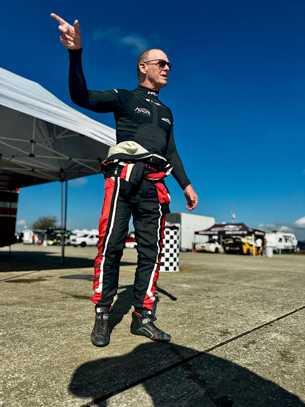 Man in racing gear gestures confidently under a blue sky. Checkered flag backdrop and vehicles visible. Energetic mood.