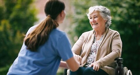 Healthcare worker in blue scrubs smiling at elderly woman in a wheelchair outdoors, surrounded by greenery. Warm, caring interaction.