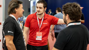 Zenon Diefenthaeler Leite, coordenador comercial da Urano Balanças e dois homens conversam em feira/conferência; o do centro usa polo vermelho Urano e crachá, em clima de networking.