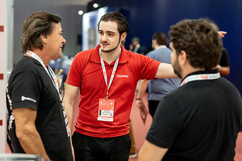 Zenon Diefenthaeler Leite, coordenador comercial da Urano Balanças e dois homens conversam em feira/conferência; o do centro usa polo vermelho Urano e crachá, em clima de networking.