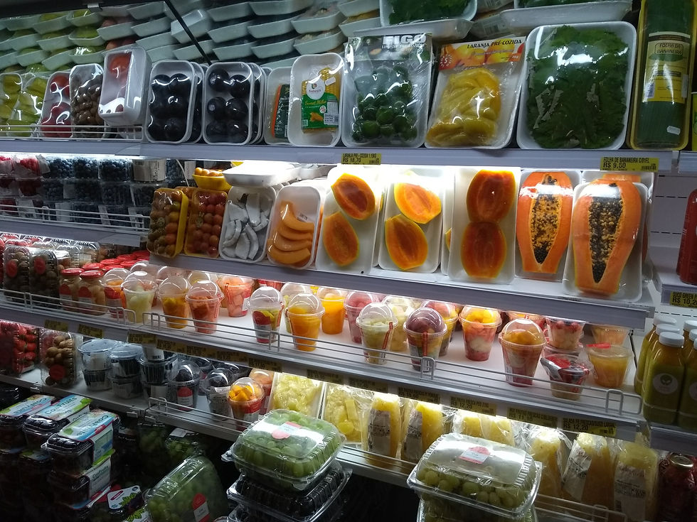 Prateleira de supermercado com frutas cortadas e sucos embalados; mangas, mamão, uvas e folhas verdes sob luz fria.