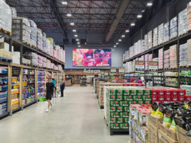 Corredor de supermercado/depósito com prateleiras altas, pessoas comprando, caixas de cerveja e a placa Adega ao fundo.