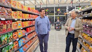 Dois homens sorrindo conversam em corredor de supermercado, com prateleiras coloridas de pães e biscoitos ao redor.