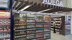 Corredor de adega em supermercado, com garrafas expostas e dois homens conversando sob a placa iluminada ADEGA.