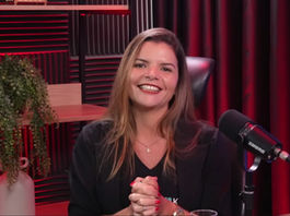 Danniela Araujo sorridente em estúdio de podcast com microfone SHURE, luzes vermelhas e plantas ao fundo, mãos juntas diante da câmera