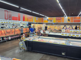 Interior de supermercado com açougue e padaria, clientes comprando carnes e pães, vitrines cheias e placas de preços.