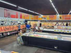 Interior de supermercado com açougue e padaria, clientes comprando carnes e pães, vitrines cheias e placas de preços.