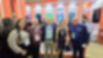 Grupo sorridente posa em estande de feira tecnológica laranja, com logos Toshiba, Mercafacil e PrompTI ao fundo.