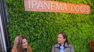 Duas mulheres conversam com xícaras de café diante de parede verde com placas MAKFRIO e IPANEMA DOCES.