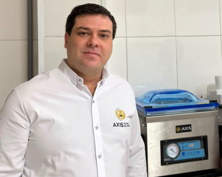 Abílio Corrêa - especialista em embaladoras e embalagens a vácuo, termoencolhimento e atmosfera modificada. É CEO e fundador da AXISVAC e vice-presidente regional da Associação Mineira de Supermercados - AMIS ao lado de uma máquina AXIS inox, em ambiente de azulejos brancos, com expressão calma.