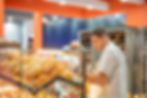 Homem escolhe pães numa padaria com vitrine de vidro, interior laranja e azul, ambiente iluminado e concentrado.