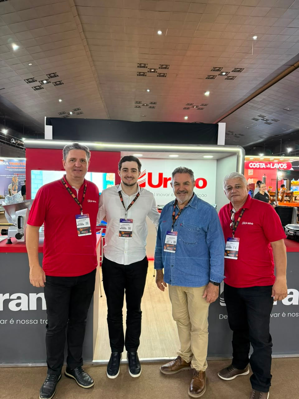 Quatro homens posam sorrindo no estande da Urano em feira, com camisetas vermelhas, camisa branca e azul, crachás visíveis.