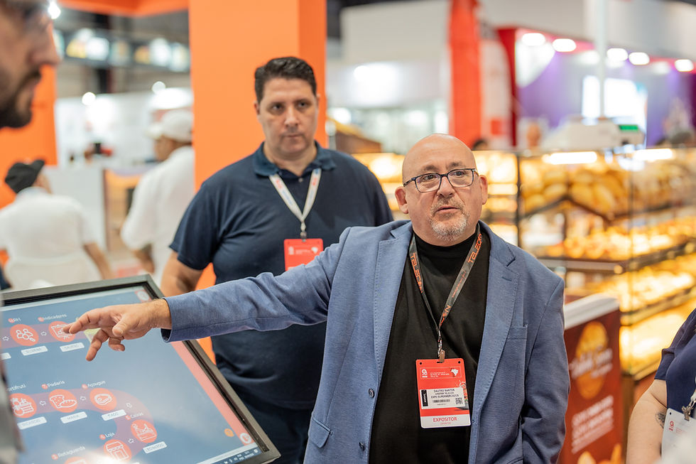 



Dautro Ribeiro CEO do Grupo Telecon aponta para tela em feira, com crachá vermelho; ao fundo, outro homem e estandes iluminados.
