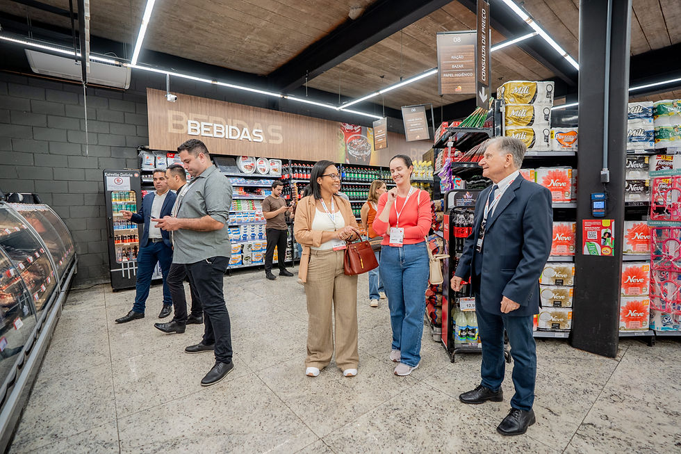 Pessoas conversam e verificam produtos em um supermercado. Placas "BEBIDAS" e "CONSULTA DE PREÇO" ao fundo. Clima descontraído.