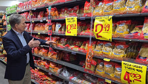 Homem de terno gesticula em corredor de supermercado, diante de prateleiras de massas e placas amarelas de preço.