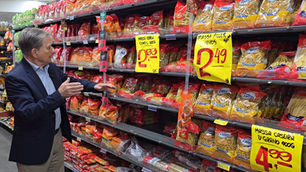 Clóvis Polese aponta para prateleira de massas em supermercado, com pacotes vermelhos e placas amarelas de preços em destaque.