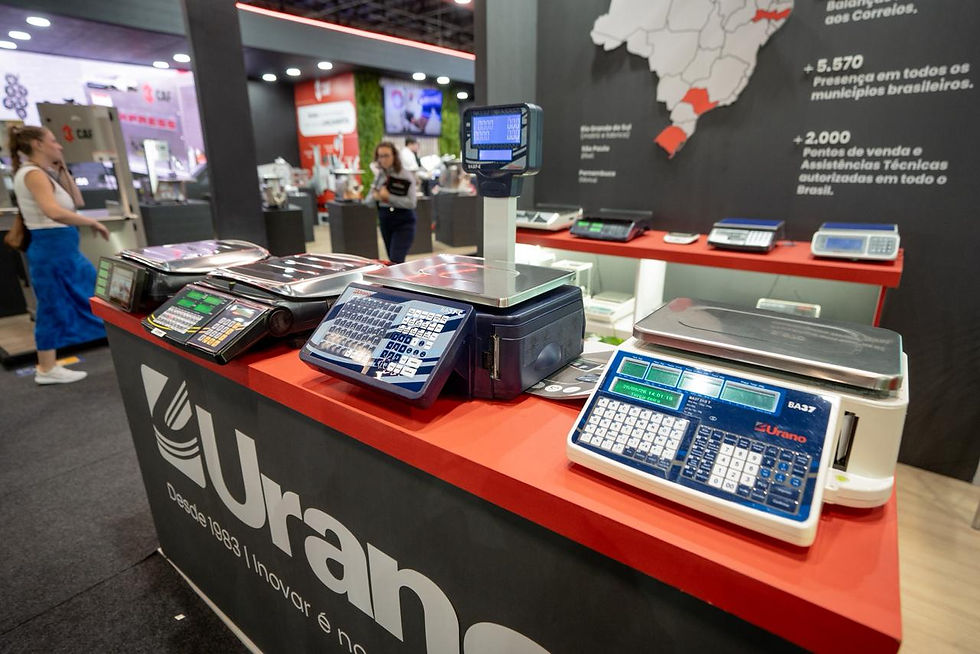 Stand da Urano com várias balanças eletrônicas sobre balcão vermelho, feira comercial ao fundo e pessoas desfocadas.