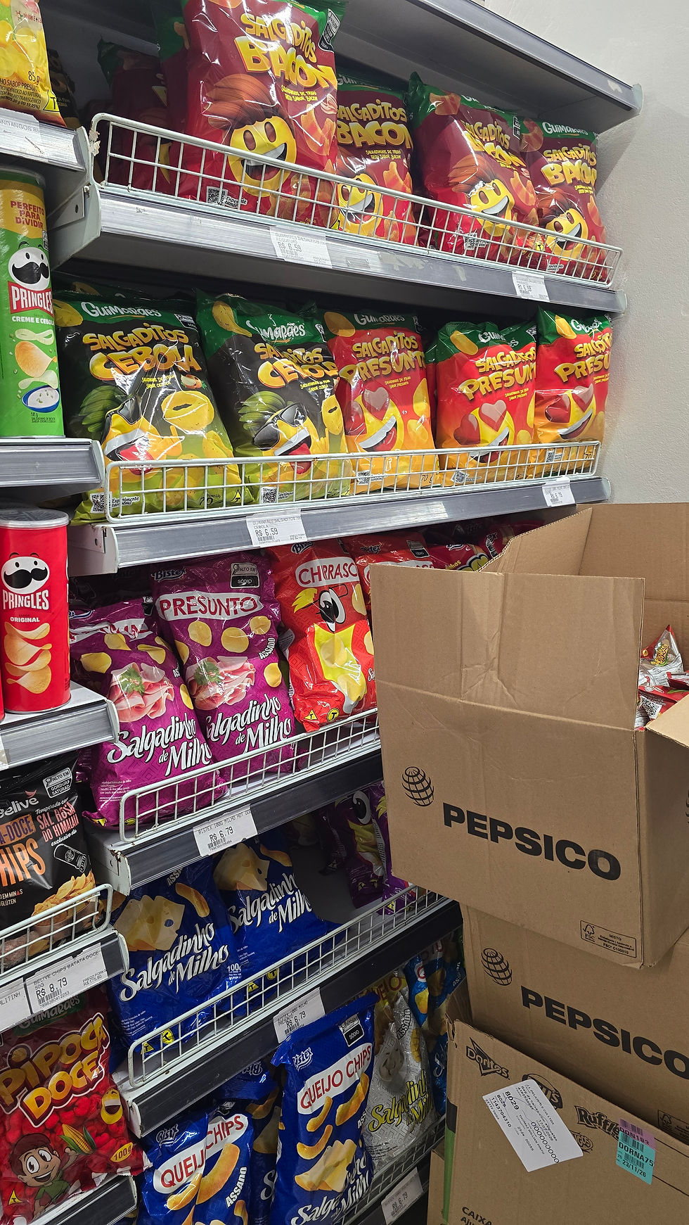 Corredor de snacks com pacotes coloridos de salgadinhos e caixas Pepsi empilhadas em prateleiras de supermercado.