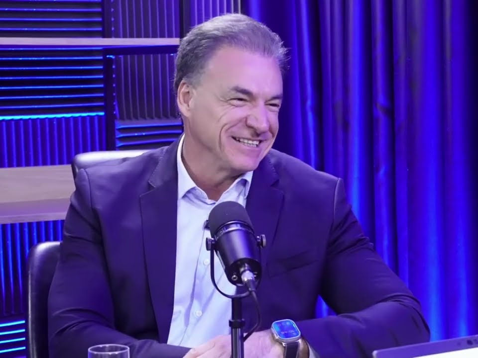 João Galassi sorrindo em estúdio de podcast roxo, falando ao microfone, de terno escuro, com fundo iluminado em azul e roxo.