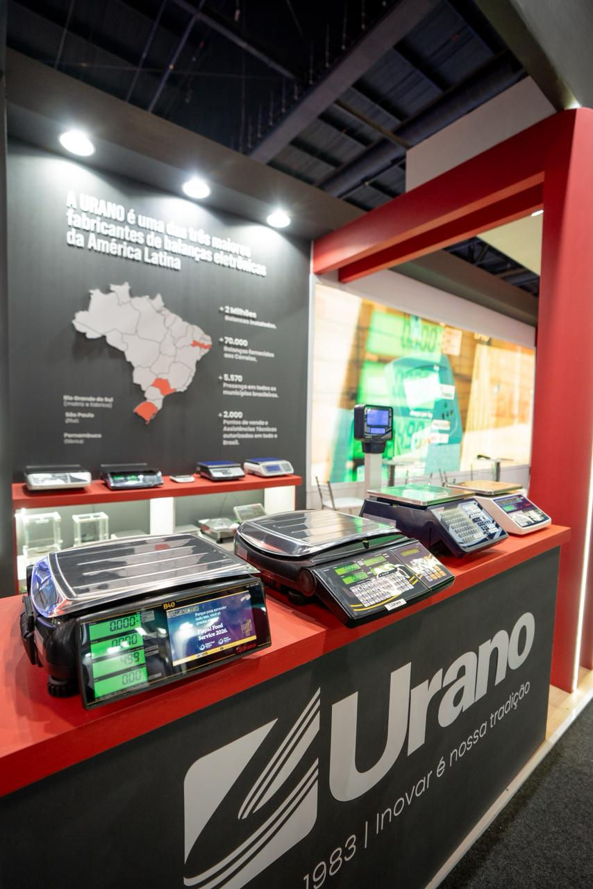 Estande da Urano com balanças eletrônicas em exposição, painel preto com mapa do Brasil e detalhes em vermelho, em feira.