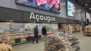 Açougue de supermercado com vitrines de carnes, clientes e promoções; pilhas de lenha à frente e letreiros de oferta.