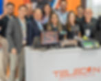Grupo de 9 pessoas sorridentes em estande da Telecon, com computadores e balança, em feira comercial.