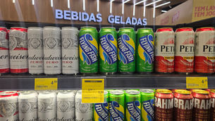 Prateleira de bebidas geladas com latas Budweiser, Flying Fish, Petra e Brahma; placas de oferta ao fundo.