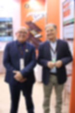 Dautro Ribeiro - CEO Grupo Telecon e Clóvis Polese - Diretor Geral da Expo Supermercados sorridentes de blazer em estande de feira, com telas e painel laranja com mão robótica ao fundo.