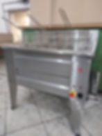 Fritadeira industrial em aço inox com cestos e botões de controle. Chão de azulejos e parede clara ao fundo. Ambiente de cozinha.