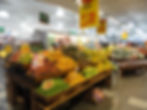 Banca de frutas em supermercado, com laranjas, maçãs, limões e abacaxis, placas de oferta em português e ambiente iluminado.
