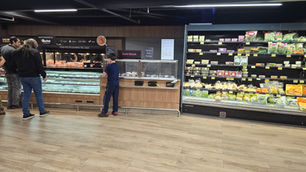 Clientes observam balcão de rotisseria e sushi em supermercado, ao lado de prateleiras de saladas e legumes.