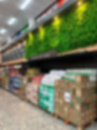 Supermercado com parede verde, letreiro Oferta do dia e pilhas de produtos em caixas e fardos no corredor.
