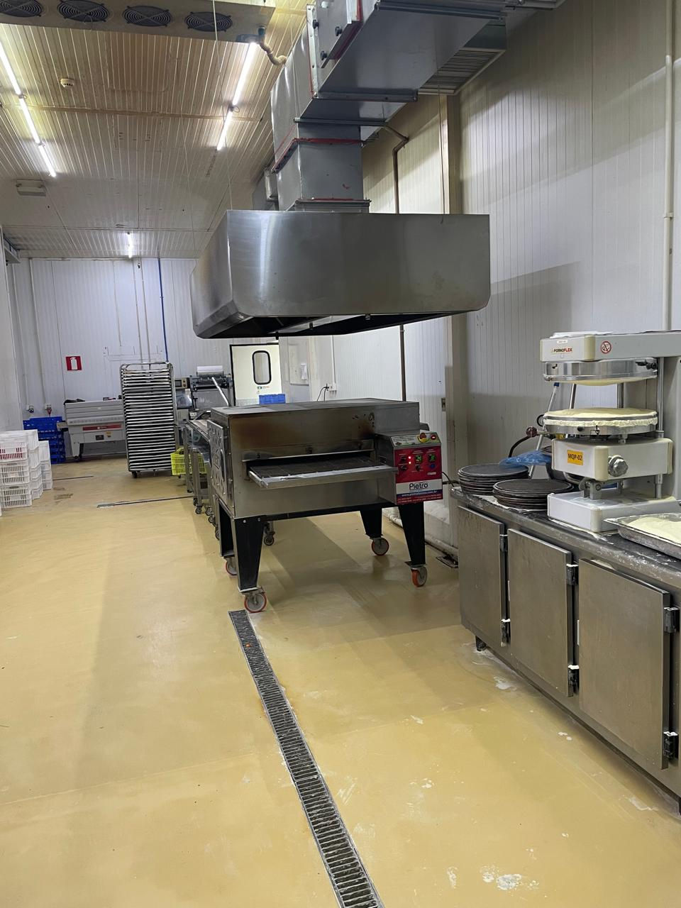 Cozinha industrial com equipamentos inox, forno grande e piso bege. Ambiente limpo e organizado, iluminação fluorescente clara.