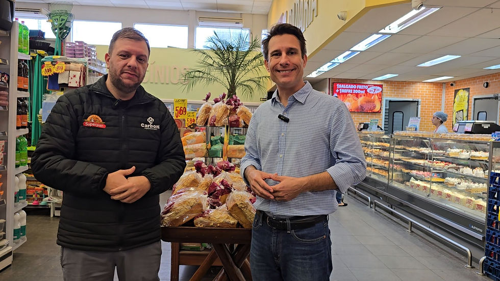 Felipe Monteiro do Carboni Supermercados e Fabiano Polese da Expo Supermercados posam sorrindo em supermercado, diante de pães embalados e vitrine de doces, com placas de preços ao fundo.