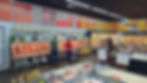 Açougue de supermercado com carnes expostas, clientes escolhendo produtos e placas de ofertas penduradas.