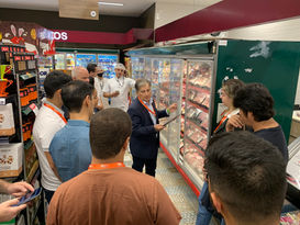 Grupo de pessoas em corredor de supermercado, ouvindo um homem de terno diante de um freezer de carnes.