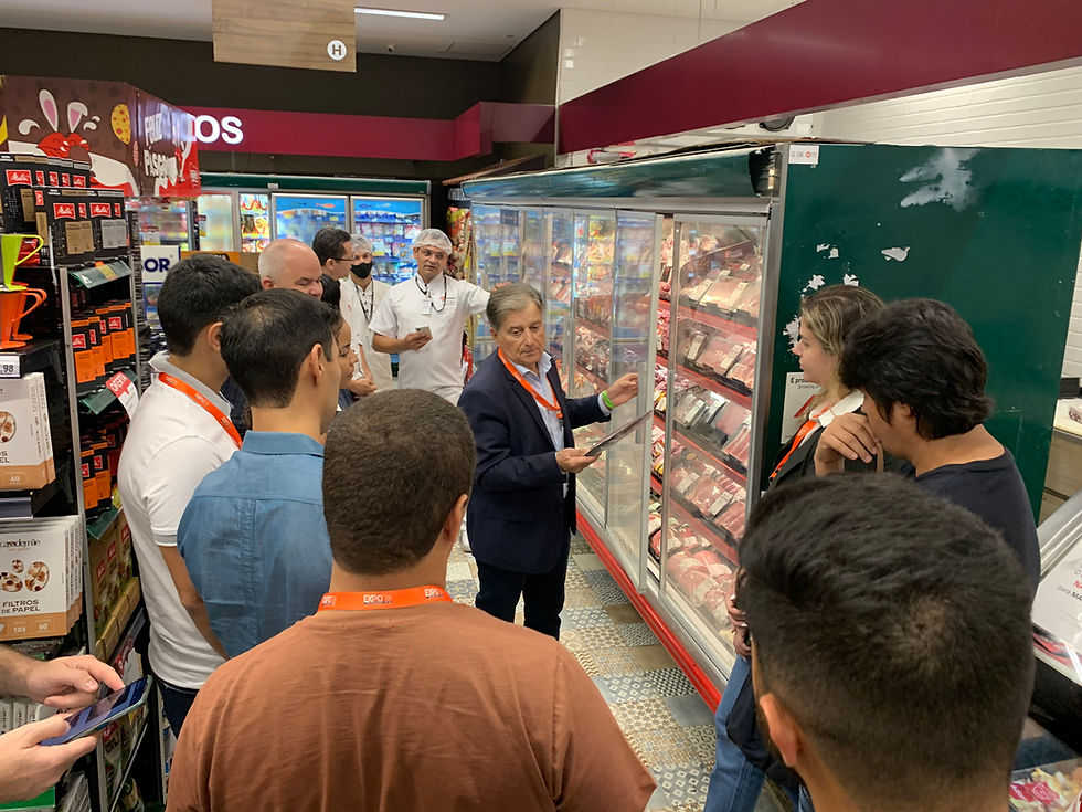 Grupo de pessoas em corredor de supermercado, ouvindo um homem de terno diante de um freezer de carnes.