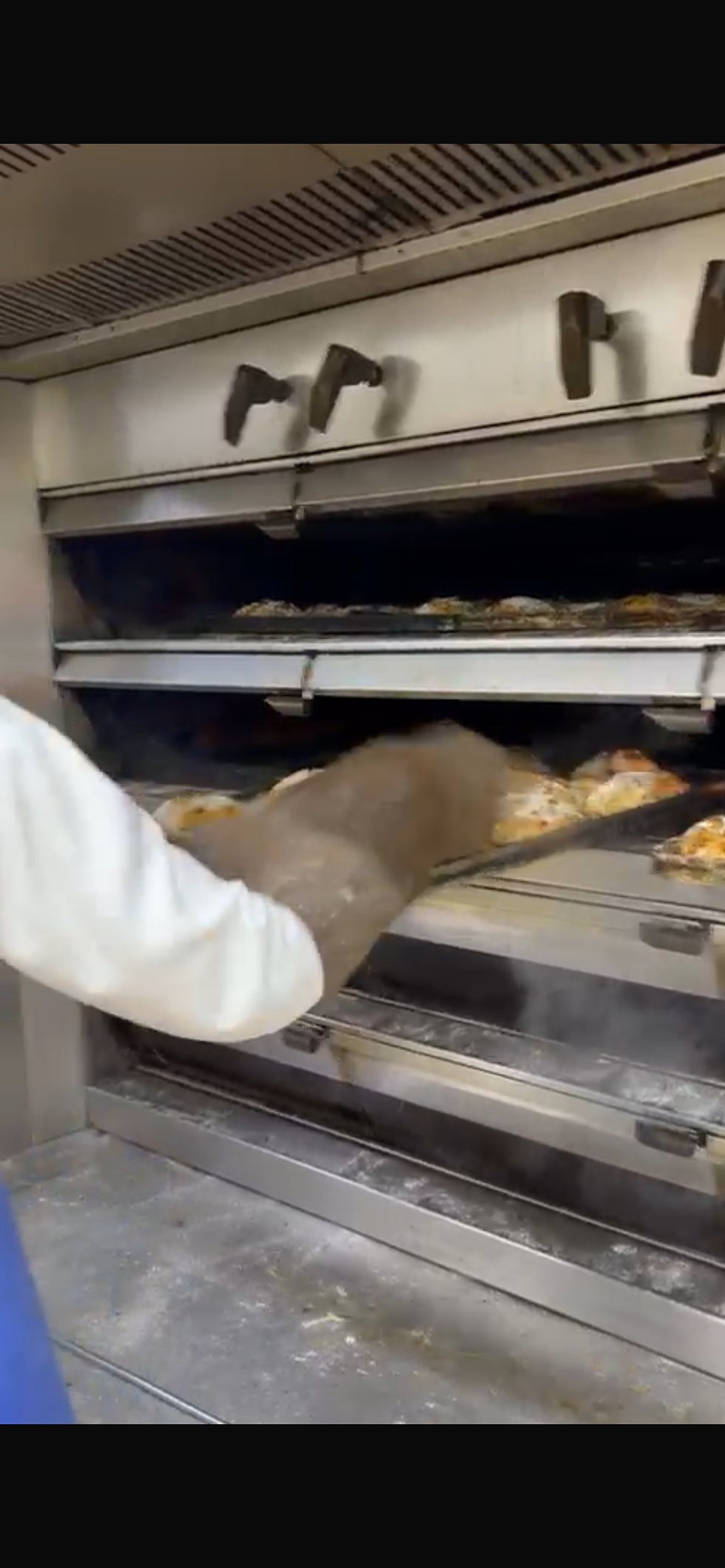 Central de produção. Padeiro retira pizzas douradas do forno de aço inoxidável, usando uma pá, em ambiente de padaria. Ação rápida e precisa.