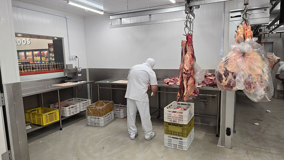 Trabalhador de branco manuseia carne em um açougue. Peças de carne penduradas, caixas plásticas e fundo com parede branca e prateleiras.