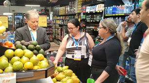 Grupo observa frutas e preços em um mercado, com prateleiras de bebidas ao fundo; clima de inspeção e conversa.