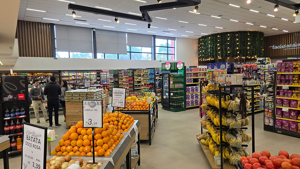 Supermercado com frutas e vegetais expostos. Três pessoas conversam. Placas de preços mostram laranjas e cebolas. Ambiente iluminado e organizado.