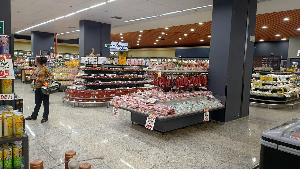 Cliente em supermercado observa seção de carnes frias e embutidos. Prateleiras cheias, teto de madeira, placas de preço visíveis.