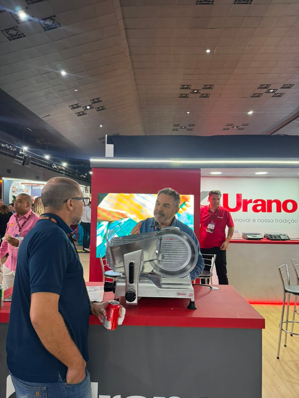 Dois homens conversam em estande da Urano numa feira, ao lado de uma fatiadora de frios; fundo vermelho e branco.