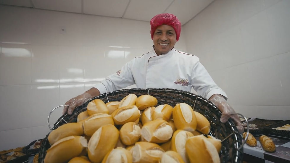 Padeiro sorridente em cozinha branca, usando touca rosa, segura grande cesta cheia de pães dourados.